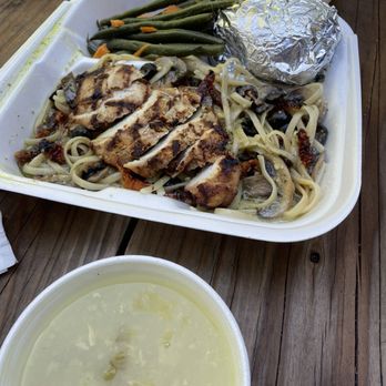 Kalamata - Mediterranean Chicken Pasta w sides of pita bread & lemon soup. IG: @bayareapineapples - The image shows a takeout meal served in a foam container placed on a wooden table. The meal includes grilled chicken slices on a bed of pasta with vegetables, a portion of green beans, and a foil-wrapped item. In front of the container, there is a cup of lemon soup with a light, clear appearance. The setting appears to be outdoors, with natural lighting and a rustic wooden surface. - Mediterranean Near Me - Arnold, California