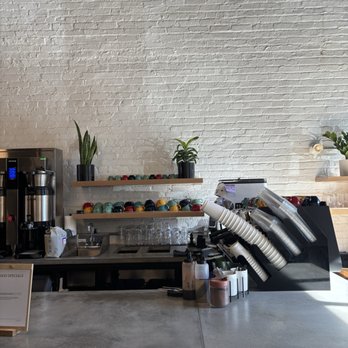 Coffee Check - The image shows a coffee shop counter with a coffee machine, various cups on shelves, and potted plants. The background consists of a white brick wall, and the counter has bottles and containers. The setting appears modern and minimalistic, with natural light illuminating the area. - Coffee & Tea Near Me - Brooklyn, New York