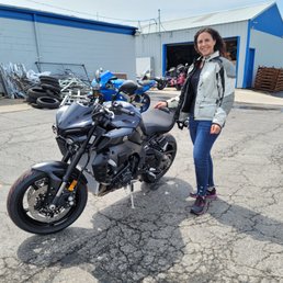 A woman wearing a motorcycle jacket and jeans stands next to a black Yamaha motorcycle in an outdoor lot. The background includes a blue and white building, motorcycle parts, tires, and other bikes. The scene appears to be at a powersport dealership specializing in Yamaha vehicles.