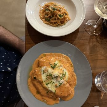 Suprema - Chicken Vodka Parmigiana - The image shows two plates of Italian cuisine on a wooden table. The larger plate contains chicken topped with a creamy orange sauce, garnished with herbs and cheese. The smaller plate holds a serving of pasta with a light sauce, garnished with herbs. There are wine glasses nearby, and the setting appears to be in a restaurant. - Italian Near Me - Rutherford, New Jersey