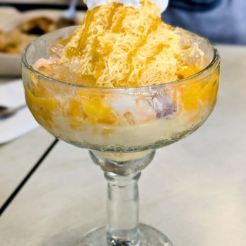 Azure - Mango bingsu - The image shows a mango bingsu served in a clear glass bowl with a stem. The dessert includes shaved ice, mango syrup, and shredded mango pieces, topped with a dollop of whipped cream. The bowl is placed on a table, with a blurred background indicating a dining setting. The presentation emphasizes the fresh mango ingredients and the icy texture of the dessert. - Desserts Near Me - Austin, Texas