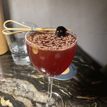Next Door - Queen for Hearts - A dark red cocktail is served in a coupe glass, garnished with a black olive on a skewer resting on the rim. The drink has a layer of small, light-colored specks on the surface. The glass is placed on a dark, patterned marble surface, with a clear glass and a black coaster visible in the background. - Speakeasies Near Me - Boston, Massachusetts