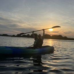 A person is seated in a kayak on a calm water body during sunset, holding a paddle above their head. The scene features a sky with clouds and the sun near the horizon, casting a warm glow. The shoreline with trees is visible in the background, and the water reflects the colors of the sky.