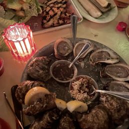 Caesar Salad - The image shows a seafood platter with several raw oysters on the half shell, served on a bed of ice with lemon wedges and dipping sauces. In the background, there are grilled chicken pieces, a salad, and a plate of sliced vegetables. The setting appears to be a restaurant or bar with dim lighting and a casual dining atmosphere.