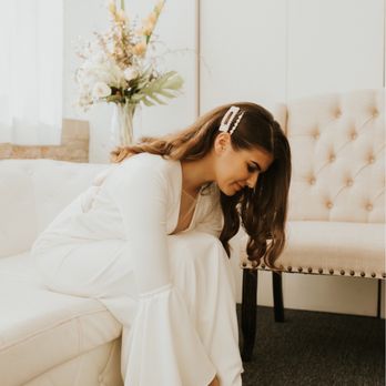 Room 1520 - Bridal suite - A woman dressed in a white gown is seated on a cream-colored sofa, leaning forward to put on beige high heels, with a floral arrangement in the background. - Venues & Event Spaces Near Me - Chicago, Illinois