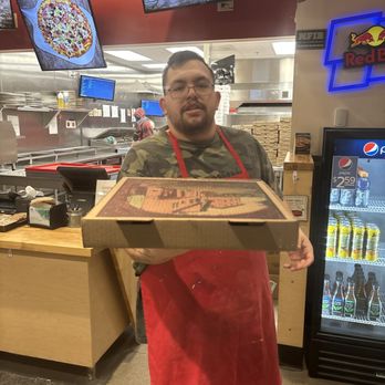 Hambone's Pizza - Service with a smile! - A man with glasses, wearing a camouflage shirt and a red apron, stands behind a counter in a pizzeria, holding a pizza box towards the camera. The background shows a kitchen area with staff, digital menu screens, and a refrigerator stocked with beverages. - Pizza Near Me - Cheyenne, Wyoming