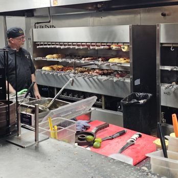 Rodizio Grill - Mesa - Open kitchen - Two kitchen staff members prepare skewered meats in a commercial kitchen, with a rotisserie grill and various utensils and ingredients on the counter. - Steakhouses Near Me - Mesa, Arizona