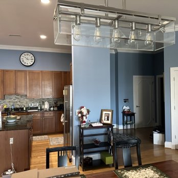 Spiro's Painting - Kitchen area - The image shows a kitchen with blue painted walls, wooden upper and lower cabinets, and a tiled backsplash behind the countertop. A clock is mounted on the wall above the cabinets. There is a refrigerator on the right side, and the countertop has various kitchen items. In the foreground, part of a dining table with chairs is visible, along with a small black shelving unit decorated with framed photos and holiday decorations. The space appears well-lit with ceiling lights. - Painters Near Me - Baltimore, Maryland