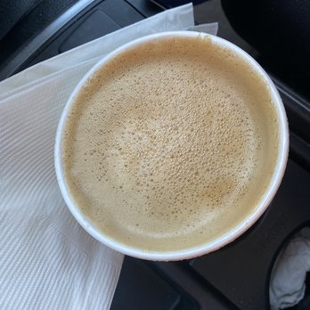 Corner Coffee Shop - Egg nog latte 16 oz - A 16 oz cup of egg nog latte with frothy foam on top is shown, held over a white napkin. The background includes part of a car interior, with a seat and a person's arm wearing a blue sleeve visible. - Coffee & Tea Near Me - Intercourse, Pennsylvania