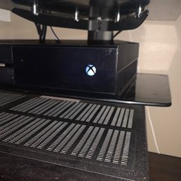 A black Xbox gaming console is positioned on a black stand, with a glowing Xbox logo visible on the front. The setup is on a surface with a textured mat underneath.