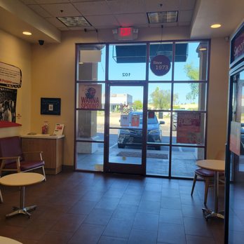 Smoothie King - Front door - The image depicts the interior of a Smoothie King store with a glass front door allowing natural light inside. There are two chairs with small round tables, a wall-mounted display, and promotional posters on the windows and walls. The store has a tiled floor and ceiling lights, with a view of parked cars and trees outside. - Juice Bars & Smoothies Near Me - Mesa, Arizona