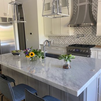 Surface Savers - Tuffskin installed by Surface Savers - The image shows a contemporary kitchen with a large marble island topped with a flower arrangement and a small potted plant. White cabinets and drawers line the walls, with stainless steel appliances including a refrigerator and a range with a decorative stainless steel hood. Two modern pendant lights hang above the island, and a patterned backsplash adds visual interest. The space appears bright and clean, with seating at the island and a neutral color palette. - Refinishing Services Near Me - Glendale, Arizona