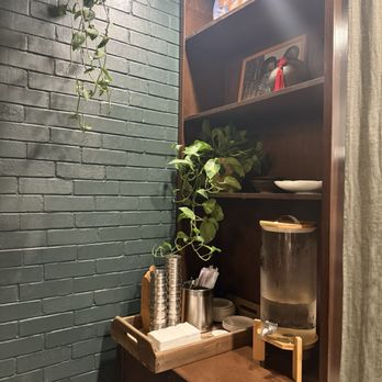 Dumpling Wei - A corner of a room features a dark green painted brick wall on the left, with a wooden shelving unit on the right. The shelves hold decorative plates, framed photos, and a potted plant. Below the shelves, a wooden tray contains disposable cups, napkins, and utensils, with a water dispenser on a small stand. A curtain is partially visible on the right side of the image. - Chinese Near Me - Pasadena, California