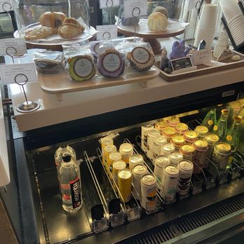 Coffee Sarap - Pastries such as croissants and bread rolls are displayed under glass covers on a wooden tray at a coffee shop. Packaged cookies are placed in front of the pastries, with labels indicating flavors. Below the display, a refrigerated section contains canned and bottled drinks, including water and juice. The setup is part of a coffee shop counter with disposable cups and other accessories visible to the side. - Coffee & Tea Near Me - Denver, Colorado