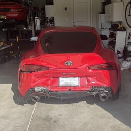 Wreck - A red Toyota Supra sports car is parked inside a residential garage, showing damage to its rear bumper. The garage contains various tools, shelves, and a bicycle mounted on the wall. A car lift with another vehicle is visible in the background, indicating ongoing vehicle maintenance or repair activities.