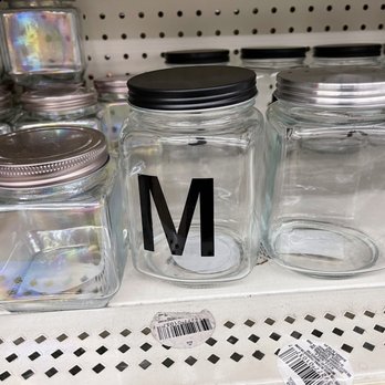 Dollar Tree - Three glass jars are placed on a store shelf, with two having metal lids and one with a black plastic lid. The jar with the letter 'M' printed on it is positioned in the middle. In the background, additional jars with metallic and holographic finishes are visible. The shelf has barcode stickers and a perforated metal surface, indicating a retail environment. - Discount Store Near Me - Placentia, California