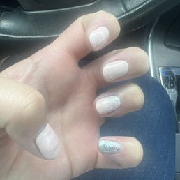 A person's hand with manicured nails, featuring a mix of plain and glittery designs, is resting on a car's steering wheel.