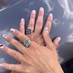 Manicures - Two hands with manicured nails featuring white and black geometric and abstract designs are resting on a reflective surface that mirrors the sky and clouds. The hands are adorned with rings, including Route 66 themed rings, and the nails have detailed nail art. The background shows a reflection of the sky with clouds, indicating the photo was taken outdoors or near a reflective surface with a view of the sky.