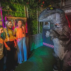 Three excited players encounter a person in an inflatable T-Rex costume, experiencing a fun scare in the Raptor Revenge room ...