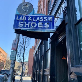Lynn Vintage Warehouse - Entrance signage - A neon sign for Lad & Lasse Shoes is mounted on the exterior of a building, with a clear blue sky and city street scene in the background. - Thrift Stores Near Me - Lynn, Massachusetts