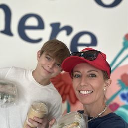 Turkey Avocado Bagel Sandwich - A woman and a boy are taking a selfie together, both smiling at the camera. The woman is wearing a red cap, sunglasses, and a chain necklace, while the boy is dressed in a white shirt. They are holding drinks and food containers, suggesting they are at a casual dining or cafe setting. The background features a colorful mural with large, stylized floral and text elements.