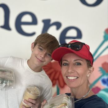 Bagel Bear - Smoothie + BLT Bagel + Turkey Avocado Bagel - A woman and a boy are taking a selfie together, both smiling at the camera. The woman is wearing a red cap, sunglasses, and a chain necklace, while the boy is dressed in a white shirt. They are holding drinks and food containers, suggesting they are at a casual dining or cafe setting. The background features a colorful mural with large, stylized floral and text elements. - Bagels Near Me - Cedar Park, Texas