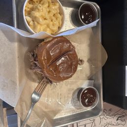 Pulled pork sandwich with mac and cheese, was very delightful. - A meal on a metal tray includes a pulled pork sandwich with barbecue sauce, a cup of macaroni and cheese, and two small cups of barbecue sauce. The sandwich is partially wrapped in paper, with a fork inserted into the meat. The macaroni and cheese appears creamy, and the sauces are dark and glossy. The tray is placed on a surface with a partially visible coloring page or menu underneath.