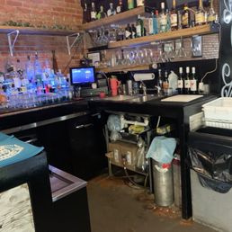 The bar - The image shows a bar counter with shelves of liquor bottles and glassware. There are two wooden shelves mounted on a brick wall, holding various bottles of alcohol and upside-down glasses. A digital cash register and a small monitor are on the counter, along with bar tools and bottles. Underneath the counter, plumbing and storage items are visible. The setting appears to be a casual bar or restaurant with a brick interior.