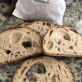 ThoroughBread - White sourdough bread had way too many holes. Crust overdone  Disappointed when I went to make sandwiches. - Three slices of sourdough bread with an open crumb structure and large air pockets are arranged on a speckled granite countertop. The slices have a golden-brown crust and a soft, airy interior. A white paper bag with a colorful 'ThoroughBread' sticker is positioned behind the slices, indicating the bakery source. The setting appears to be a kitchen or bakery workspace. - Bakeries Near Me - Austin, Texas