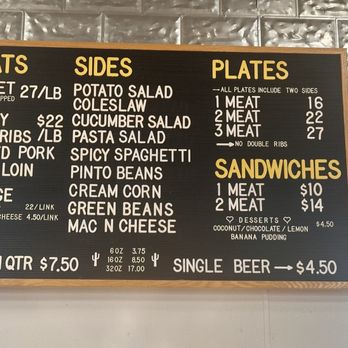 Lum's - Inside Menu - A rectangular black menu board with a wooden frame lists various food items available at a barbecue restaurant. The menu is divided into sections for meats, sides, plates, sandwiches, and drinks, with prices and portion sizes included. The meats section features options like brisket, turkey, pork ribs, pulled pork, pork loin, and sausage. Sides include potato salad, coleslaw, cucumber salad, pasta salad, spicy spaghetti, pinto beans, green beans, and mac n cheese. Plates come with two sides, and sandwiches are available with one or two meats. A single beer is also listed. The menu uses a mix of uppercase and lowercase text, with some items highlighted in yellow. - Barbeque Near Me - Junction, Texas