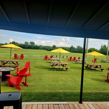 19th Tee Golf Range - Beautiful. Haven't been here in 40 years! - A grassy outdoor area features red Adirondack chairs, picnic tables, and yellow umbrellas, with a clear sky and trees in the background. - Sporting Goods Near Me - Ottawa, Ontario