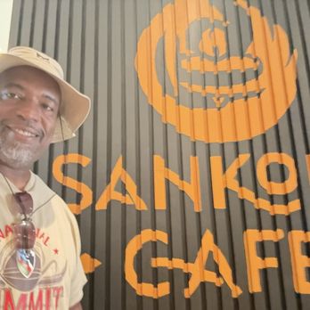 Sankofa Cafe - A man with a hat and sunglasses poses in front of a black wall with orange Sankofa Cafe signage. - Coffee & Tea Near Me - Montclair, New Jersey