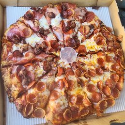 Half pepperoni/half peaks - A large pizza in a cardboard box features a variety of toppings including pepperoni, sausage, ham, and melted cheese. The pizza is cut into slices, with some toppings overlapping the edges of the slices. The box is open, revealing the pizza inside, and the pizza appears freshly baked with a golden crust.