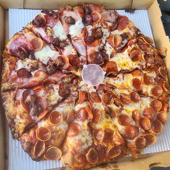 Mountain Mike's Pizza - Half pepperoni/half peaks - A large pizza in a cardboard box features a variety of toppings including pepperoni, sausage, ham, and melted cheese. The pizza is cut into slices, with some toppings overlapping the edges of the slices. The box is open, revealing the pizza inside, and the pizza appears freshly baked with a golden crust. - Pizza Near Me - Orem, Utah
