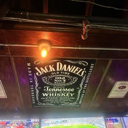 A Jack Daniel's whiskey sign is affixed to a dark wooden ceiling, featuring branding and product information. Two Tito's Handmade Vodka signs are positioned on either side of the Jack Daniel's sign. The ceiling includes visible pipes and a light bulb, indicating an indoor bar setting.