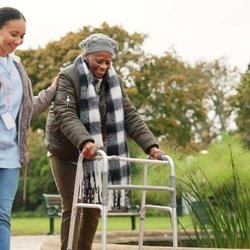 SYNERGY HomeCare - A caregiver supporting a senior woman using a walker during an outdoor walk, highlighting SYNERGY HomeCare's focus on helping...