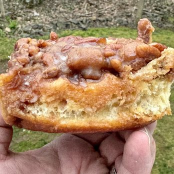 Flour Bakery + Cafe - sticky sticky bun - A close-up image shows a sticky bun with a golden-brown crust, topped with caramel glaze and chopped nuts. The bun is held in a person's hand, with the background featuring grass, soil, and trees, indicating an outdoor setting. The bun appears freshly baked, with a soft interior and a glossy, sticky topping. - Coffee & Tea Near Me - Cambridge, Massachusetts