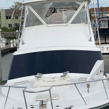 Big Boy Wraps - Ocean Yacht - A white motorboat with a dark blue stripe along the hull is moored at a marina. The boat has a clear windshield, metal railings, and various equipment on the deck, including a winch and cleats. In the background, there are residential buildings with multiple stories and a cloudy sky. The marina is equipped with docks and mooring posts, and other boats are visible nearby. - Vehicle Wraps Near Me - Martinez, California