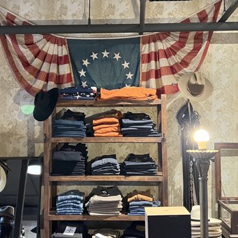 Cave & Post Trading - Lots of American Made choices. - A wooden shelving unit displays neatly folded men's clothing, including jeans, t-shirts, and pants in different colors such as orange, black, white, and blue. Behind the shelf, a large American flag with a circle of stars is draped on the wall. Hats and accessories are hung on the side of the shelf, and a vintage-style lamp with a globe bulb is positioned nearby. The setting appears to be inside a retail store specializing in men's fashion and accessories. - Men's Clothing Near Me - Phoenix, Arizona