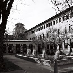 The image depicts a black-and-white photograph of a multi-story building with numerous windows, situated behind a small garden area with ornamental grasses and a fountain. Leafless trees frame the scene, and a sidewalk runs along the front of the building, suggesting a winter or early spring setting.