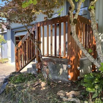 Domnich Construction - New railing - A wooden staircase with a railing ascends to a house entrance, with trees and greenery around it, and a house number visible. - Handyman Near Me - Eugene, Oregon