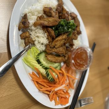 Wrap N Roll - Chicken and Rice plate - The image shows an oval white plate with a serving of cooked chicken pieces placed on a bed of white rice. The plate also includes sliced cucumbers, shredded carrots, and a small plastic container of dipping sauce. The dish is set on a wooden table, with a fork and chopsticks nearby, indicating a meal at a restaurant or casual dining setting. - Vietnamese Near Me - Albuquerque, New Mexico