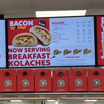 Shipley Do-Nuts - The image shows a digital menu board at Shipley Do-Nuts in Leander, TX, promoting breakfast kolaches. The menu lists different types of kolaches such as sausage, ham & cheese, egg & sausage, and egg & bacon, with prices and calorie information. The board features images of the kolaches and emphasizes that they are handmade daily. Below the menu, red Shipley Do-Nuts boxes are displayed on a shelf, and part of an interior space with a ceiling, lighting, and an exit sign is visible. - Donuts Near Me - Leander, Texas