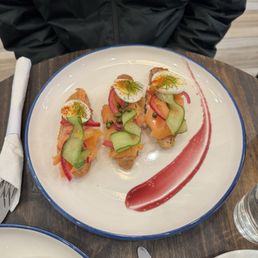 Smoked Salmon Tartine - Three smoked salmon tartines are presented on a white plate with a blue rim. Each tartine features a slice of bread topped with smoked salmon, a halved boiled egg garnished with herbs, and thinly sliced cucumber and pickled red onion. A red sauce smear decorates the plate's side. The plate is placed on a dark wooden table, with a fork, knife, and napkin nearby, and a person wearing a black jacket is seated behind the plate.