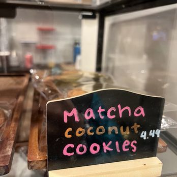 Caphe Den - pastry - A black sign with pink, orange, and white handwritten text advertises matcha coconut cookies priced at $4.49. The sign is placed on a wooden stand inside a display case, with baked goods visible behind the glass. The background shows a kitchen or bakery setting with shelves and containers. - Coffee & Tea Near Me - Flushing, New York