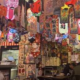 Dinner - The image shows the interior of a restaurant decorated with numerous hanging paper lanterns in various colors, including pink, red, yellow, and white, each with Japanese characters and designs. The walls are covered with posters and artwork, many featuring Japanese text, traditional imagery, and anime characters. There are tables set with soy sauce bottles, and a few patrons are seated, enjoying their meals. The overall ambiance is vibrant and themed around Japanese culture.