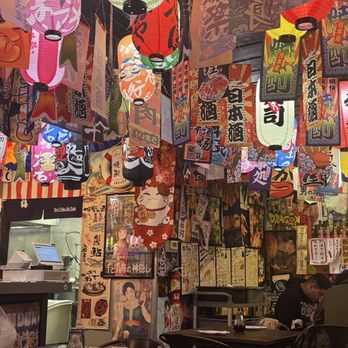 Dragon Sushi - Dinner - The image shows the interior of a restaurant decorated with numerous hanging paper lanterns in various colors, including pink, red, yellow, and white, each with Japanese characters and designs. The walls are covered with posters and artwork, many featuring Japanese text, traditional imagery, and anime characters. There are tables set with soy sauce bottles, and a few patrons are seated, enjoying their meals. The overall ambiance is vibrant and themed around Japanese culture. - Sushi Bars Near Me - Albuquerque, New Mexico