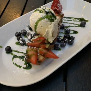 Roost - A dessert is presented on a white oval plate, including a scoop of vanilla ice cream topped with green sauce, fresh sliced strawberries, and whole blueberries. The plate is garnished with additional blueberries, green sauce streaks, and a dusting of powdered sugar, set on a dark wooden table. - New American Near Me - Sparkill, New York