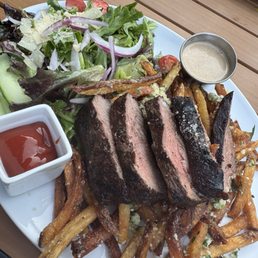Steak Frites - A plate of sliced steak frites with a side salad containing cherry tomatoes, lettuce, and onions. The dish includes a small cup of sauce and a ramekin of dipping sauce, all served on a white plate placed on a wooden table. The fries are scattered around the steak and salad, with some topped with grated cheese or seasoning.
