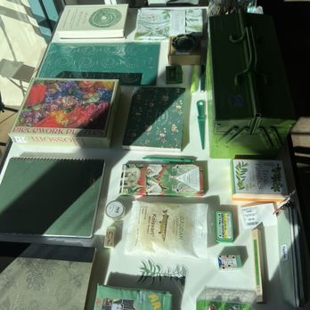 Noted. - Stationary - The image shows a table filled with stationery and gift items, including notebooks, puzzle boxes, and decorative objects. Sunlight creates shadows across the items, highlighting their textures and colors. The table also features a green metal box, a glass jar, and a potted plant in the background, suggesting a retail or display setting for stationery and home goods. - Cards & Stationery Near Me - Ojai, California